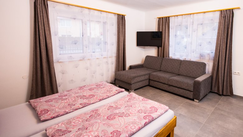 Ein Schlafzimmer mit Doppelbett, grauem Sofa, Fernseher und zwei Fenstern mit Vorhängen.