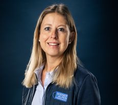Portraitfoto Wiener Alpen Mitarbeiterin, die eine Jacke mit Wiener Alpen Logo trägt und vor einem dunklem Hintergrund steht.
