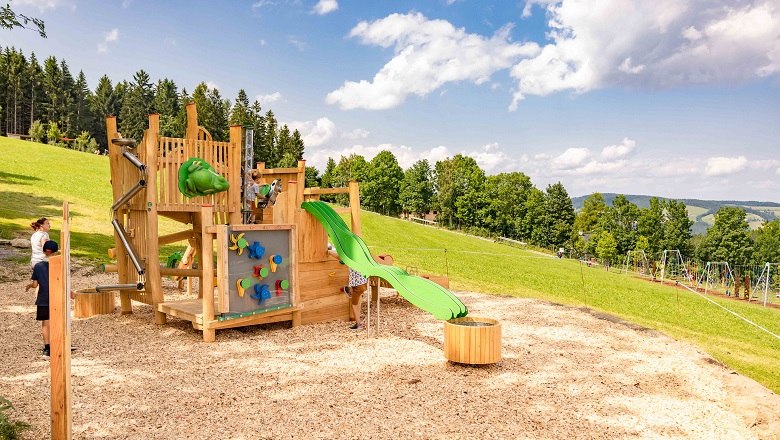 Spielplatz mit Klettergerüst und Rutsche auf einer Wiese, umgeben von Bäumen und blauem Himmel.