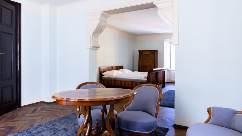 Ein elegantes Zimmer mit antiken Möbeln, darunter ein Holztisch, zwei Stühle und ein Bett im Hintergrund.