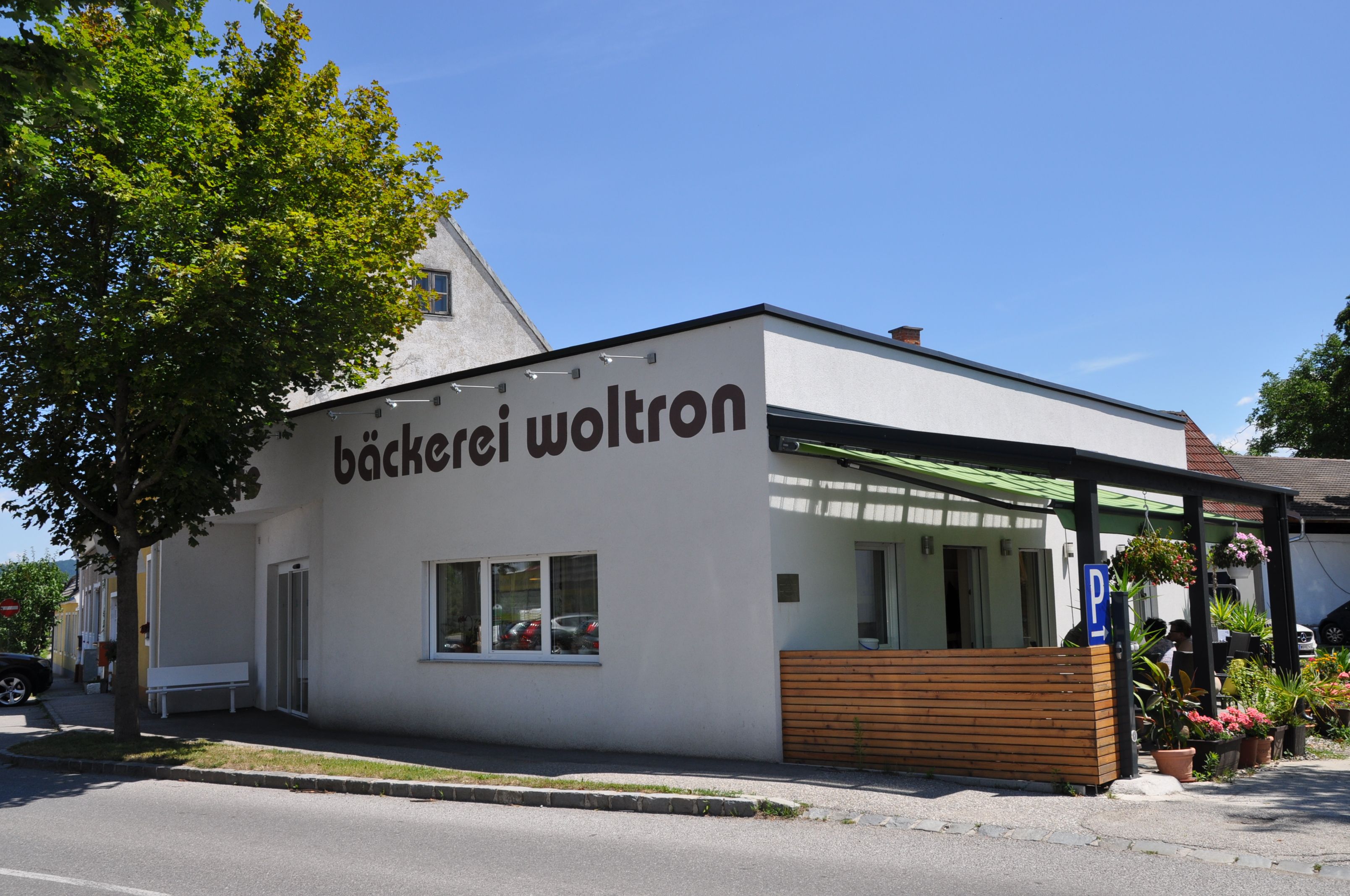 Außenansicht der Bäckerei Woltron mit Pflanzen und Parkplätzen.