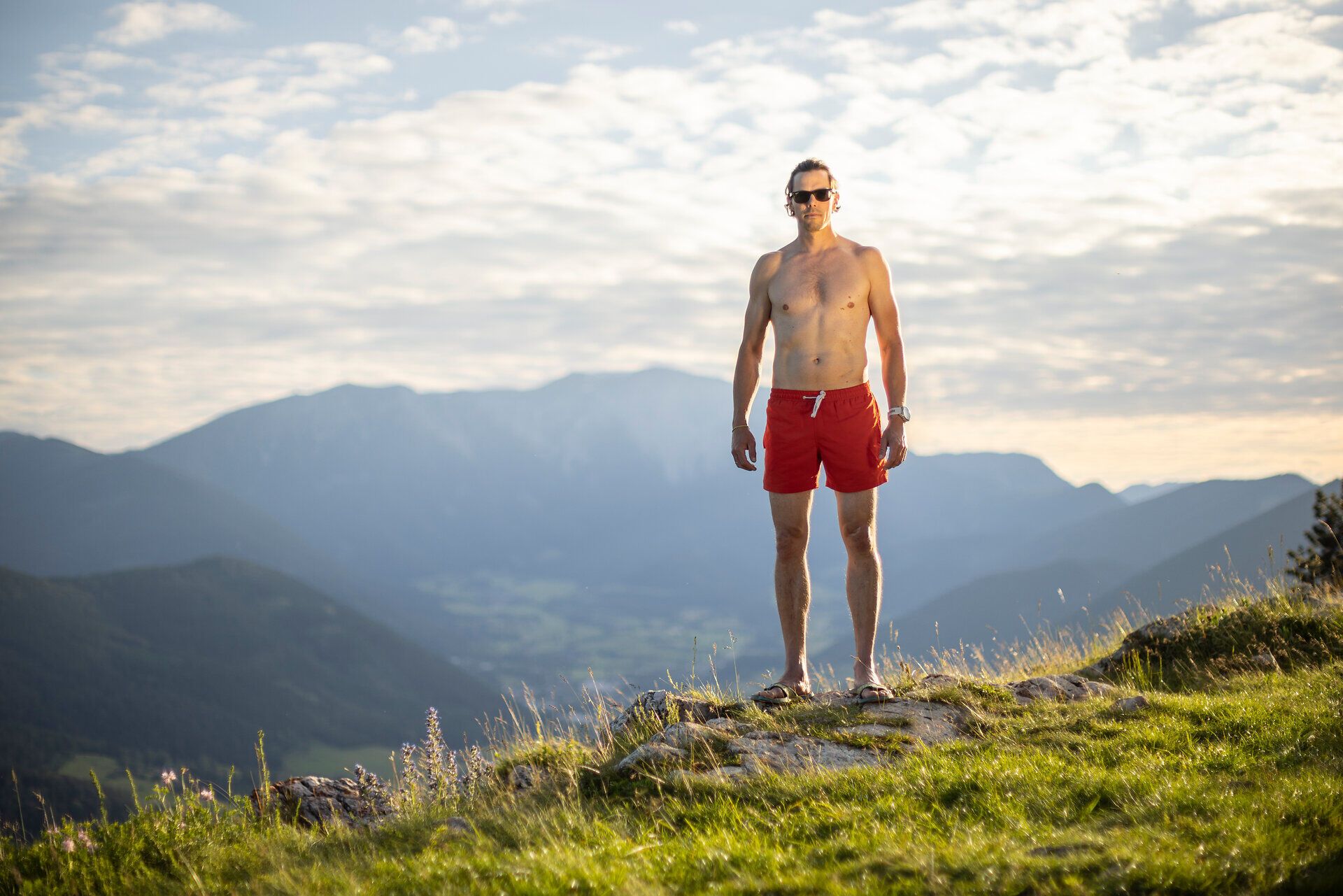 Mann mit Sonnenbrille und roten Shorts steht auf einem grasbewachsenen Bergkamm, im Hintergrund sind Berge und ein wolkiger Himmel zu sehen.