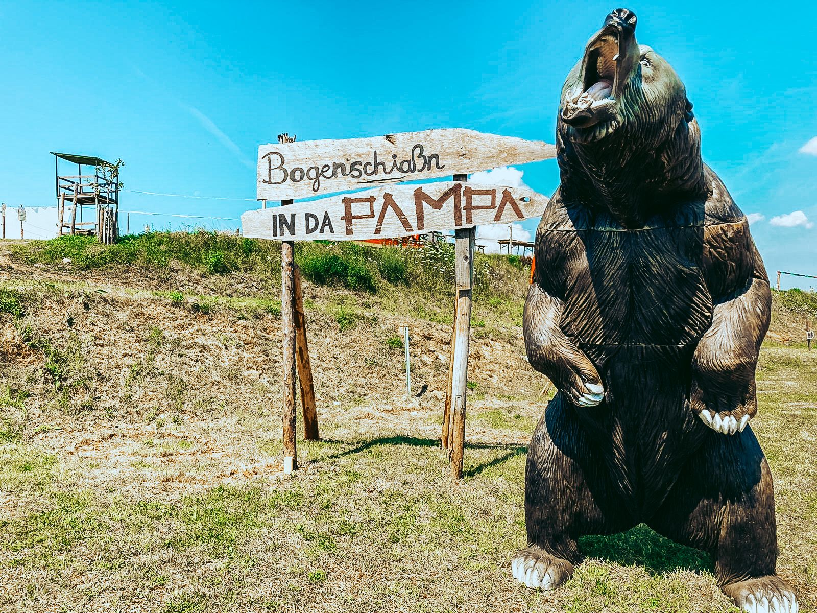 A large bear model next to a sign with the inscription 'Bogenschiaßn in da Pampa' on a meadow.