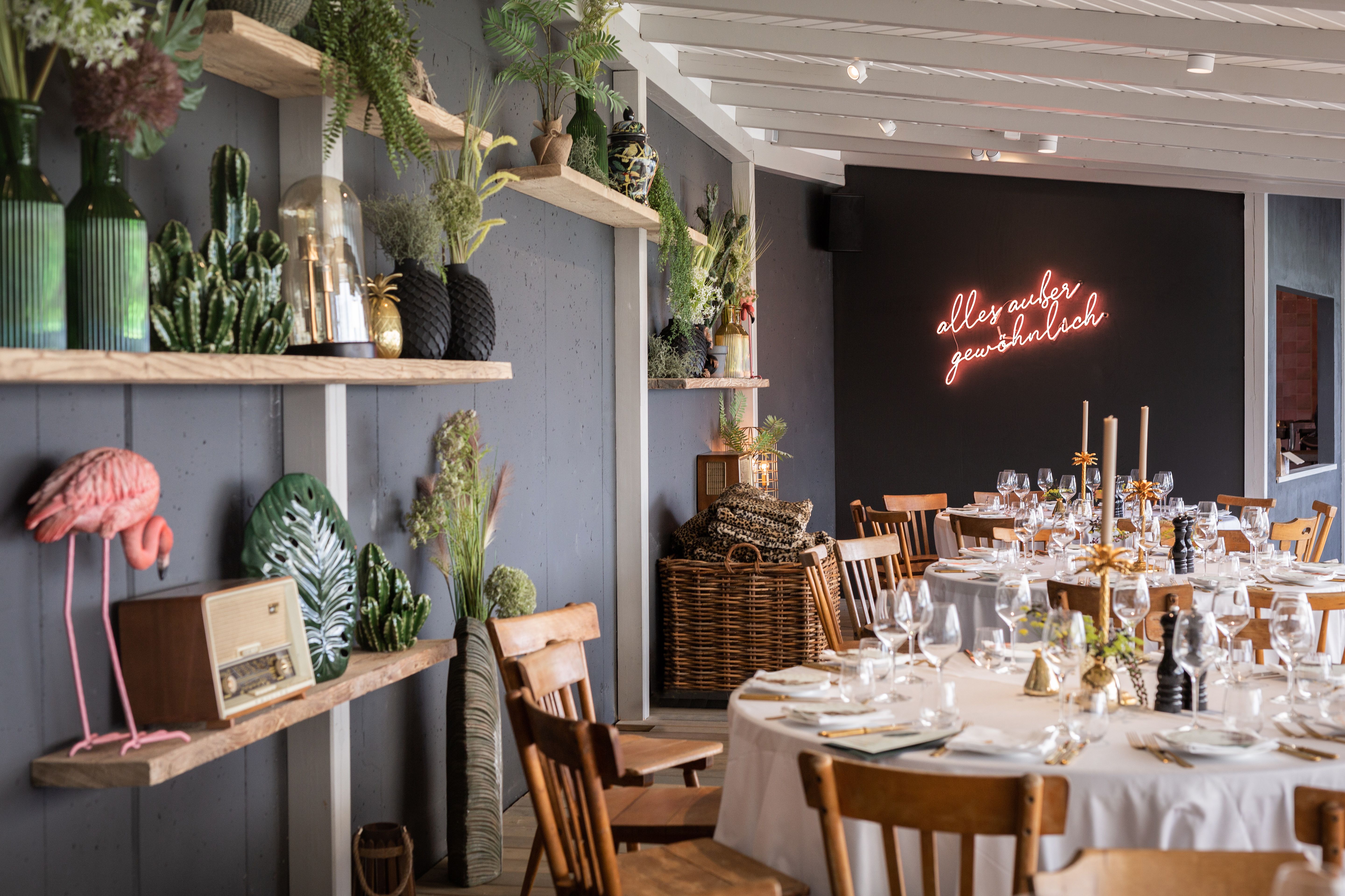 Ein stilvoll dekorierter Raum mit gedeckten Tischen, Pflanzen und einem Neon-Schriftzug an der Wand.
