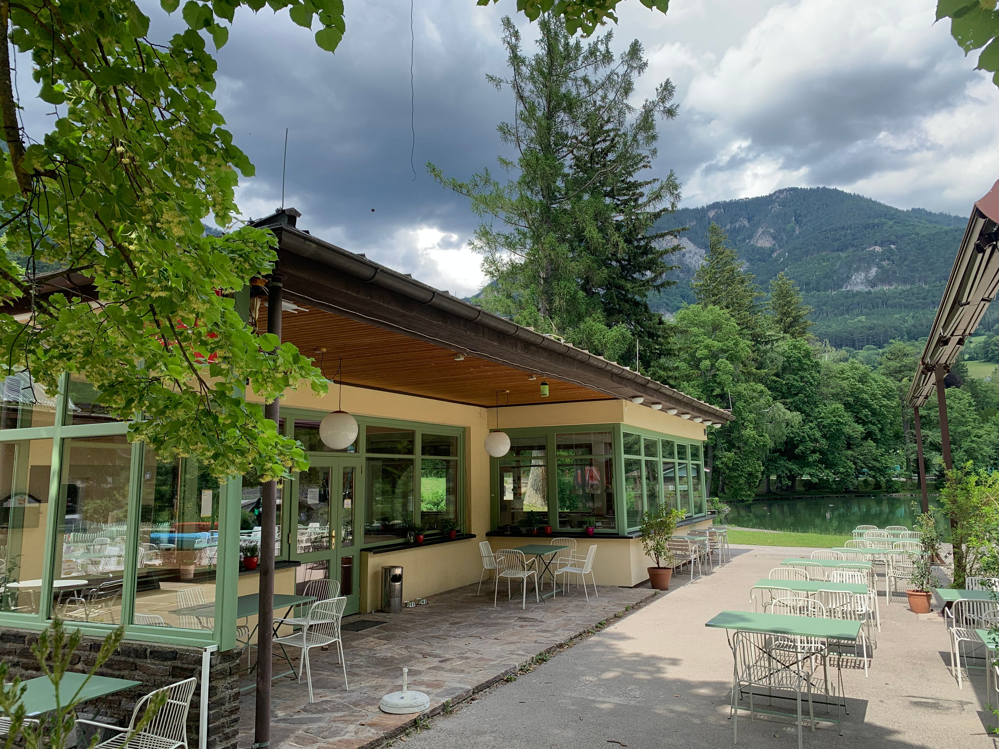 Ein Café mit Terrasse, umgeben von Bäumen und Bergen, unter einem bewölkten Himmel.