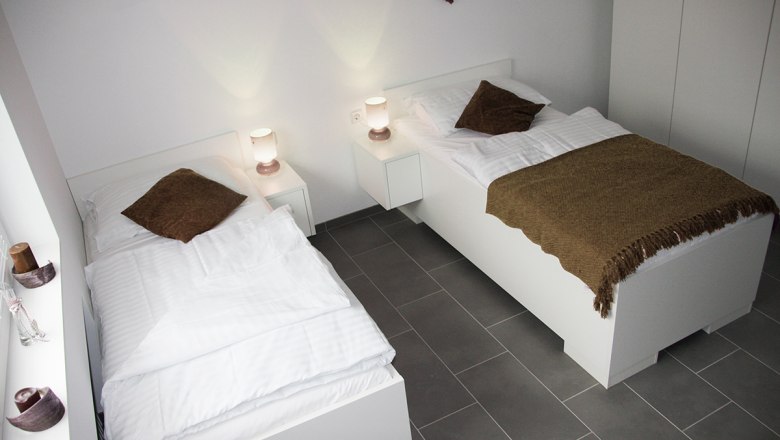 Zwei Einzelbetten in einem modernen, hellen Zimmer mit weißen Möbeln und braunen Decken.