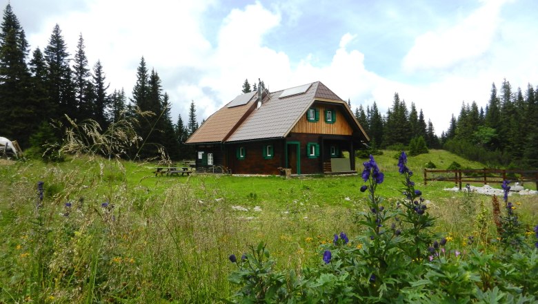 Eine Holzhütte in einer grünen Wiese mit Blumen und Bäumen im Hintergrund.