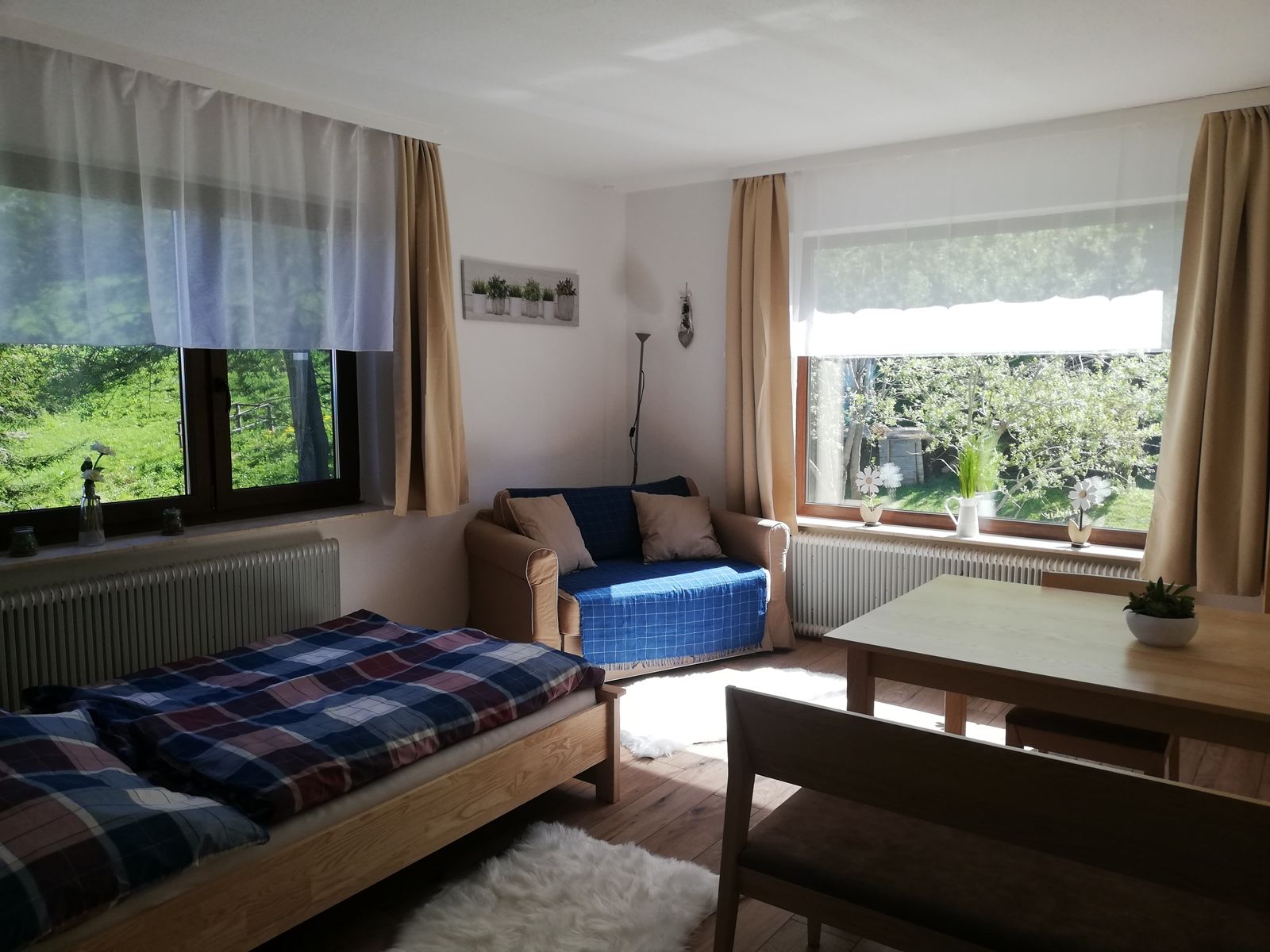 Helles Wohnzimmer mit Bett, Sofa, Tisch und großen Fenstern mit Blick ins Grüne.