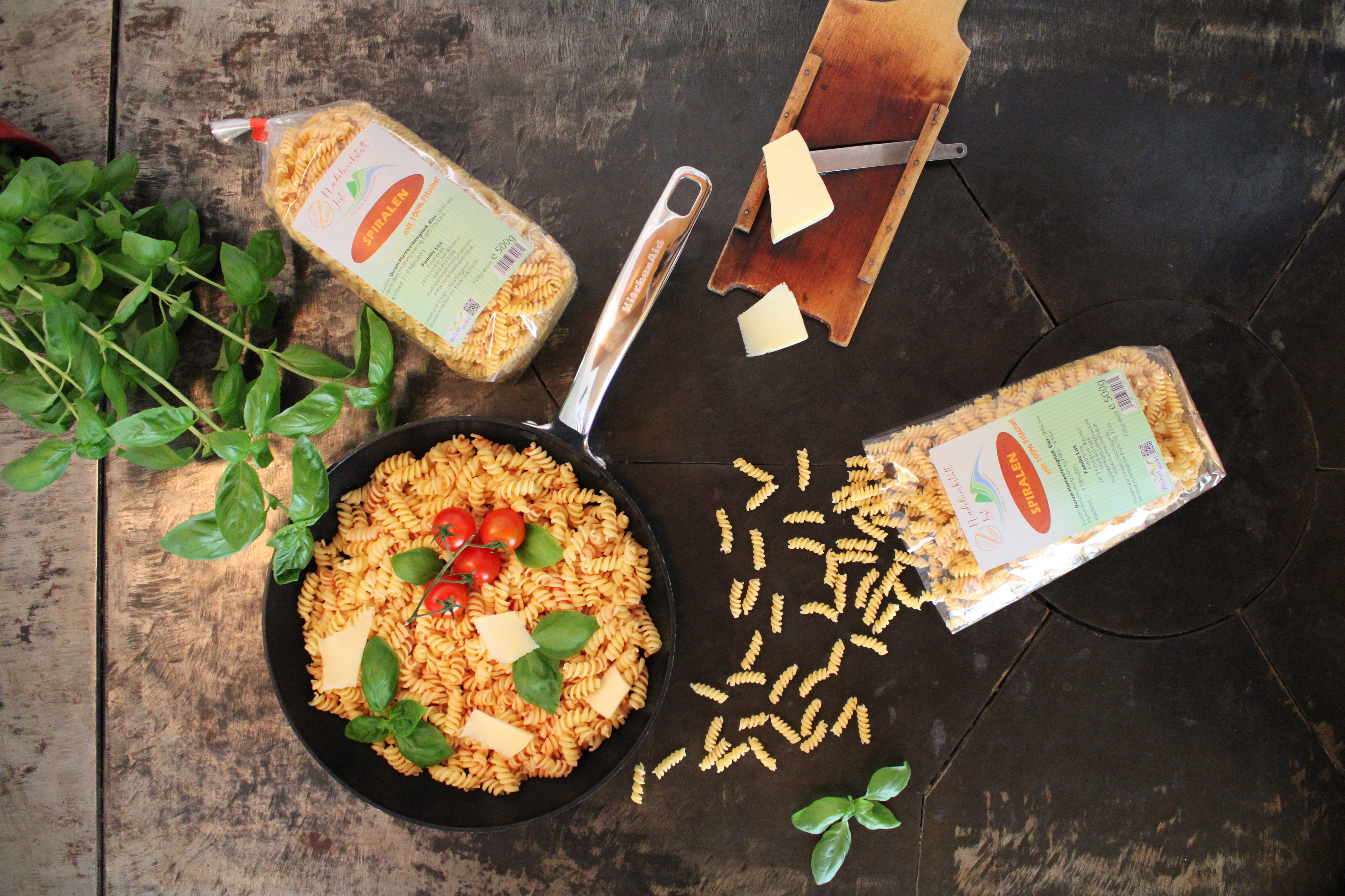 Eine Pfanne mit Spiralnudeln, Tomaten und Basiikum auf einem Holztisch, umgeben von Nudelpackungen, Basilikum und Käse.