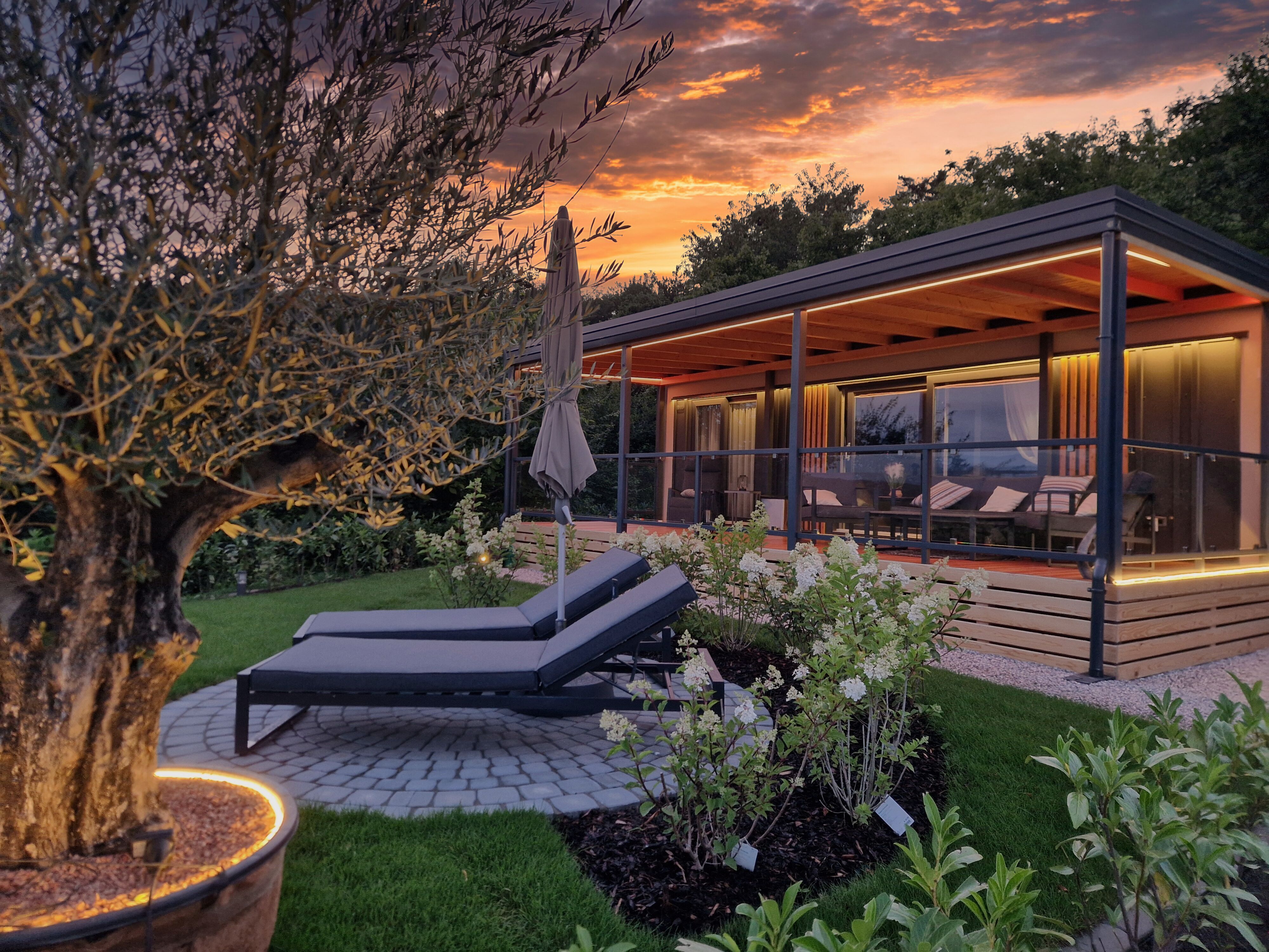 Ein modernes Chalet mit Terrasse und Liegestühlen im Garten bei Sonnenuntergang.