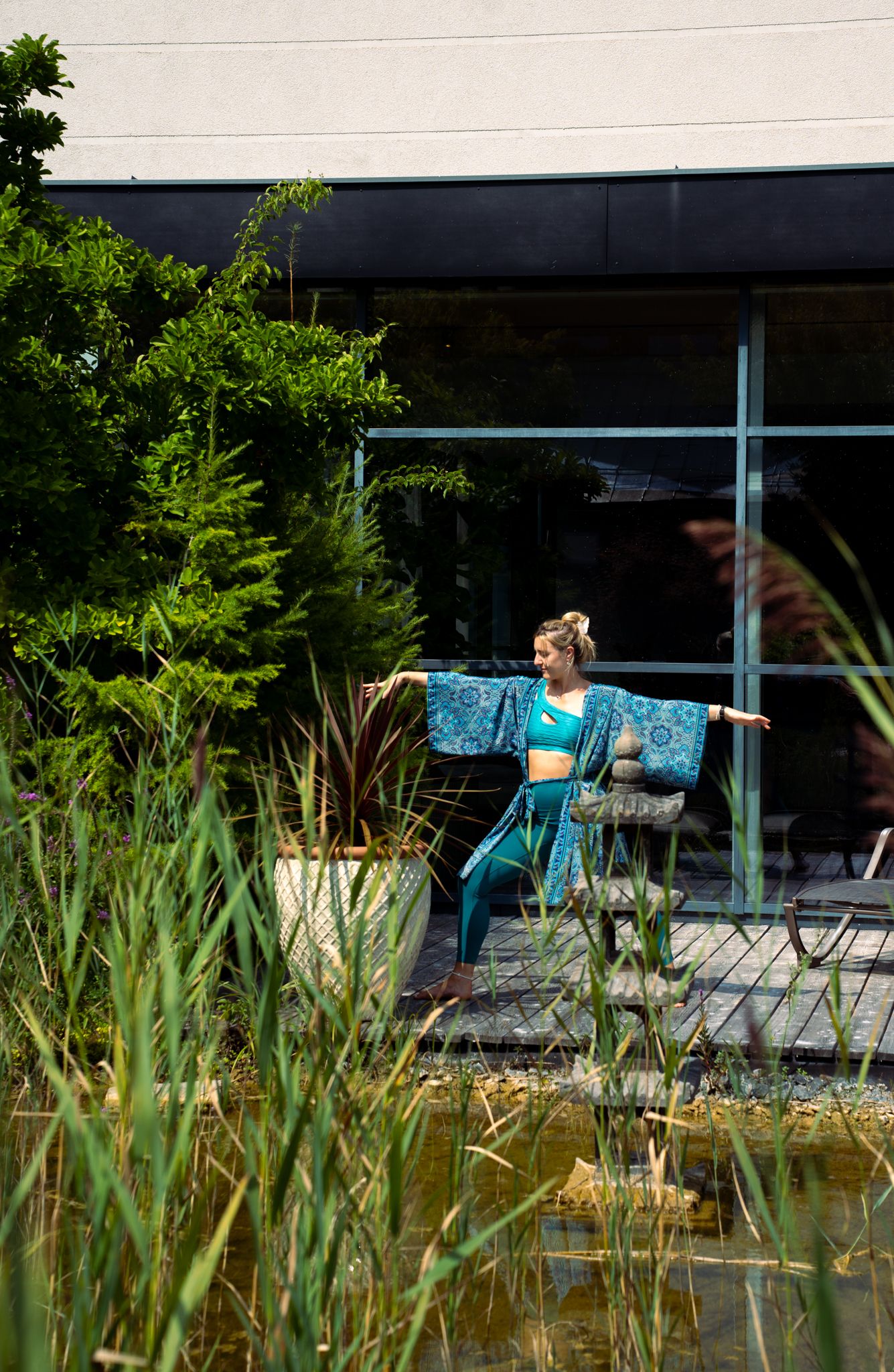 Eine Frau in blauer Yogakleidung macht eine Yoga-Pose im Freien neben einem Teich und Pflanzen.
