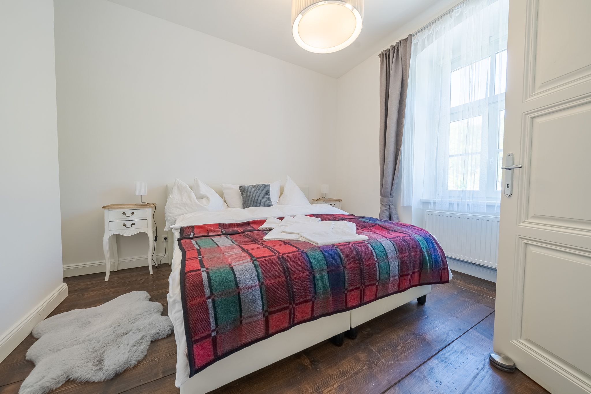 Ein gemütliches Schlafzimmer mit einem Doppelbett, das mit einer bunten Decke bedeckt ist. Ein kleiner Nachttisch steht daneben, und ein Fenster lässt Tageslicht herein.