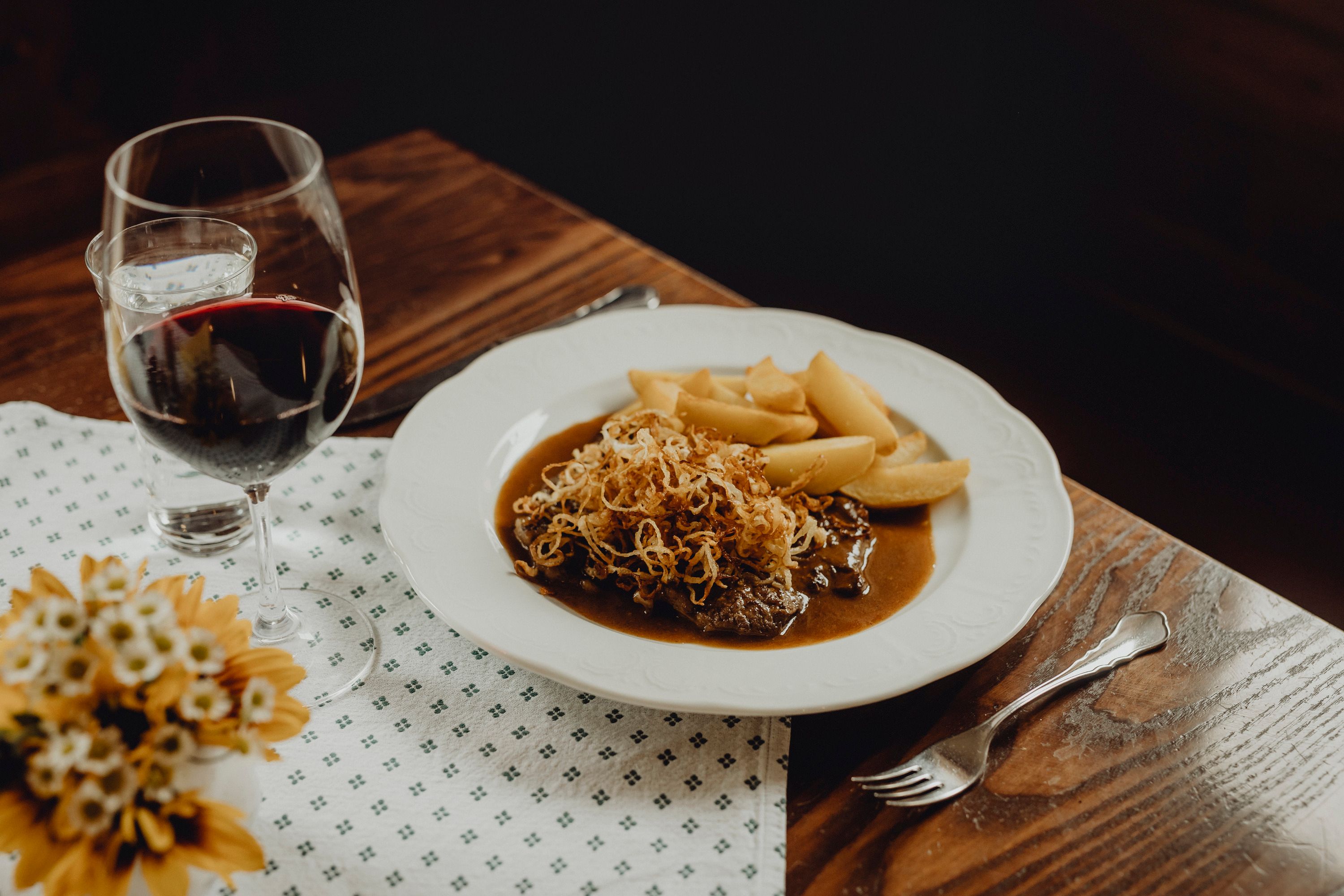 Ein Teller mit Zwiebelrostbraten, Kartoffeln und einem Glas Rotwein auf einem Holztisch.