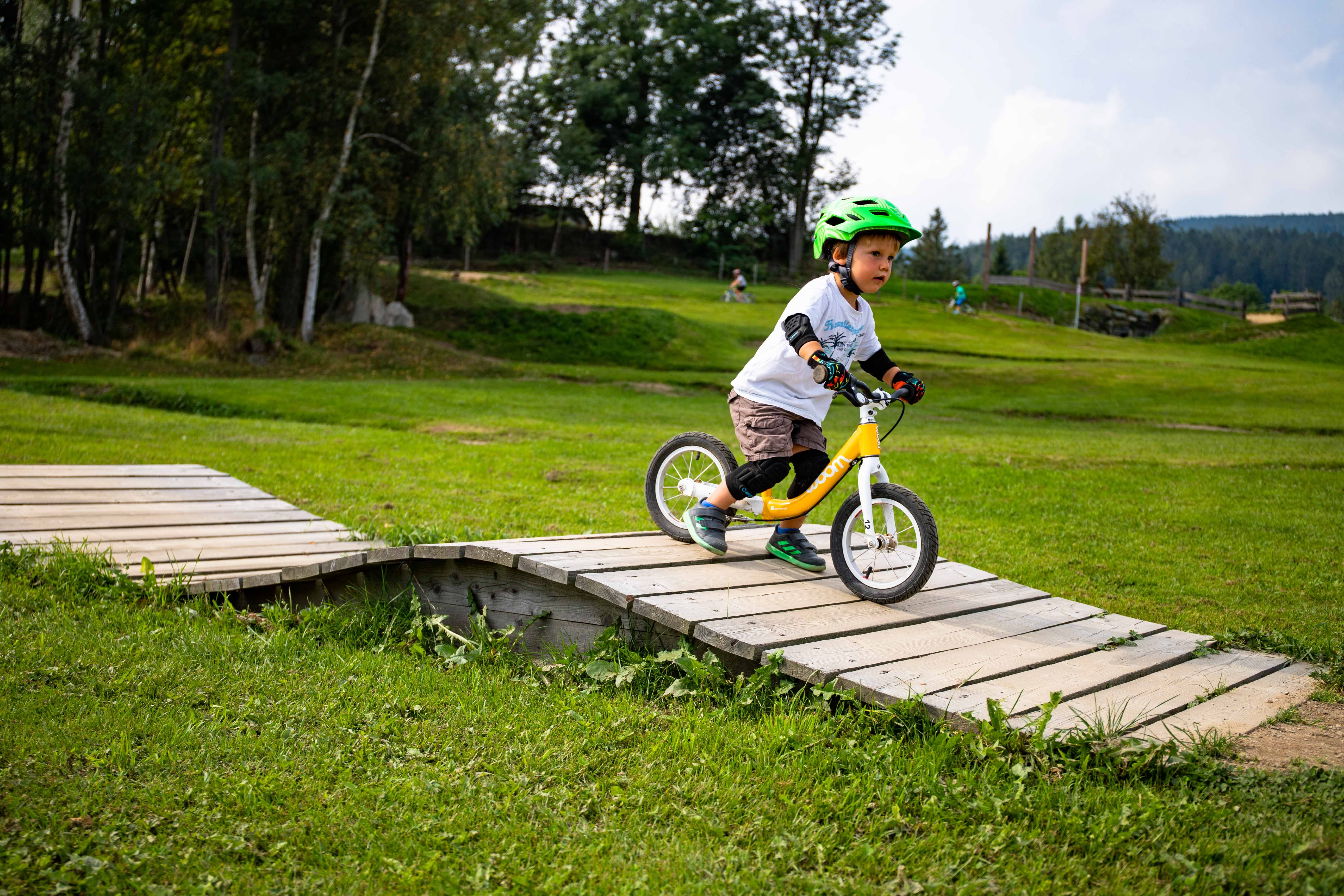 Ein Kind mit grünem Helm fährt auf einem Laufrad über eine Holzrampe im Mini Bikepark Wexl Trails.