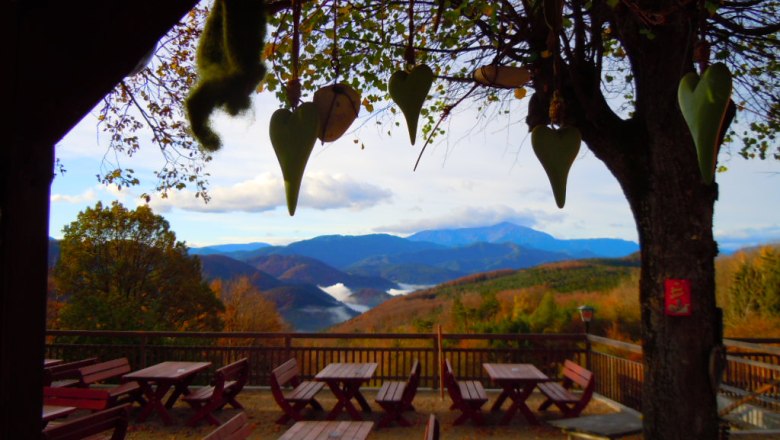 Terrace with wooden tables and benches, surrounded by trees, with a view of wooded mountains and clouds on the horizon.