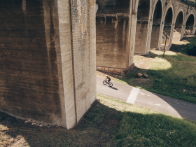 Ein Radfahrer gleitet entspannt auf einem schmalen Weg, umgeben von der majest&auml;tischen Kulisse der Wiener Alpen. Die sanften H&uuml;gel und die beeindruckenden Br&uuml;cken schaffen eine harmonische Verbindung zwischen Natur und Architektur, die zum Verweilen einl&auml;dt.