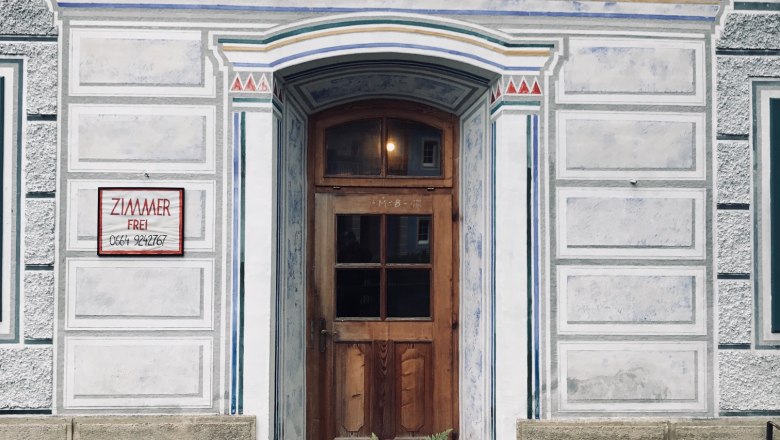 Entrance to an ornate building with a wooden door and decorative paintings, surrounded by a wooden fence and ferns.