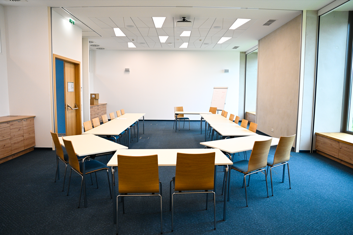 Großer, heller Raum mit blauem Teppichboden, Tageslicht, Stühlen und Tischen.