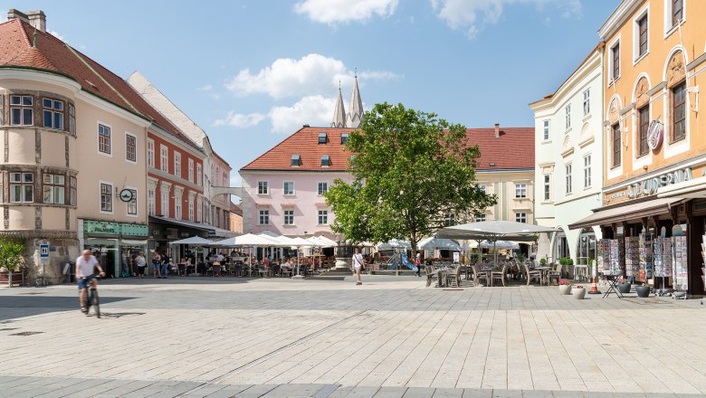 A lively main square with caf&eacute;s and stores, surrounded by historic buildings. People sit outside, a cyclist rides past. Blue sky with clouds.