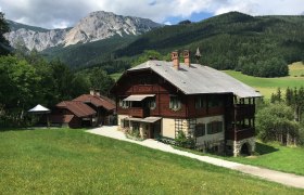 Ein traditionelles Holzhaus in einer bergigen Landschaft mit grünen Wiesen und bewaldeten Hügeln im Hintergrund.
