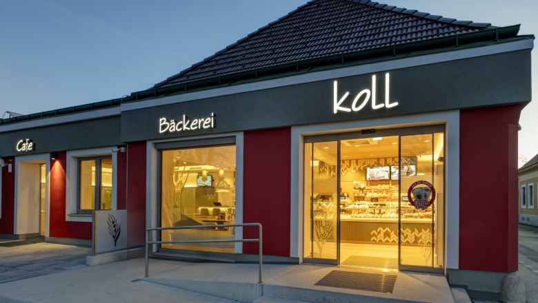 Außenansicht am Abend der Bäckerei mit beleuchtetem Schild 'Bäckerei Koll' und 'Cafe'.