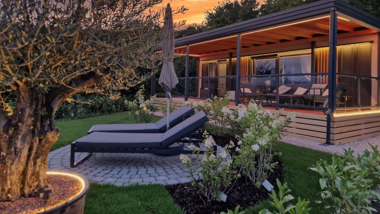 Ein modernes Chalet mit Terrasse und Liegestühlen im Garten bei Sonnenuntergang.