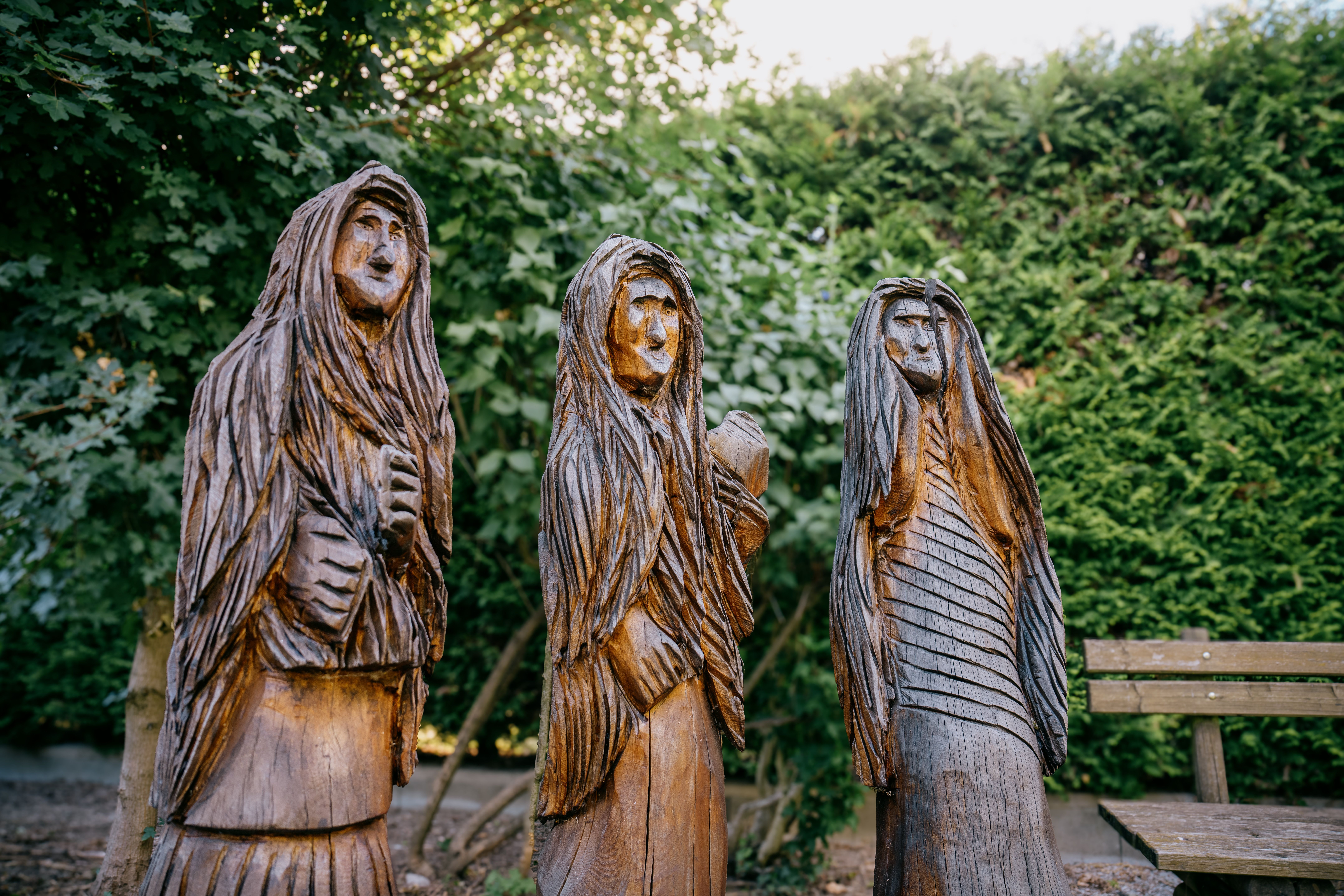 Drei geschnitzte Holzfiguren von Hexen am Sagenweg Lanzenkirchen vor dichtem Grün und einer Bank.