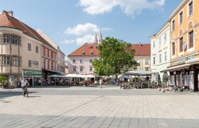 A lively main square with cafés and stores, surrounded by historic buildings. People sit outside, a cyclist rides past. Blue sky with clouds.