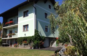 Ein grünes Haus mit Balkonen voller Blumen, umgeben von Bäumen und einem gepflegten Garten.