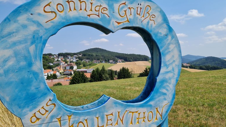 Blauer Holzrahmen in Form einer Wolke mit Schriftzug 'Sonnige Grüße aus Hollenthon' vor Landschaft.