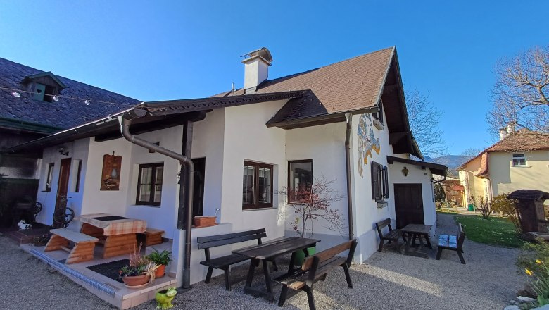 Leo-Hof, © Wiener Alpen Ein traditionelles Haus mit Holzbänken und Tischen im Garten, umgeben von blühenden Pflanzen und Bäumen unter einem klaren blauen Himmel.