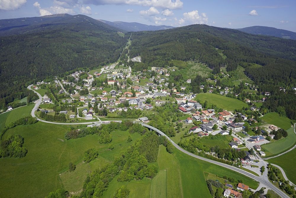 Luftaufnahme eines Dorfes in einer grünen, hügeligen Landschaft mit Wäldern und Wiesen.