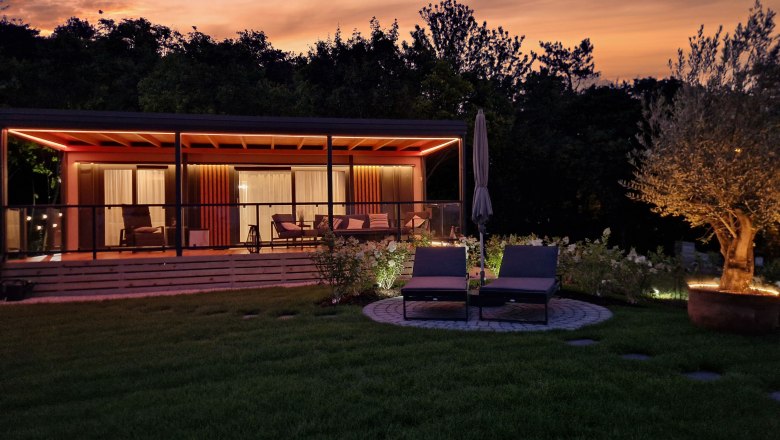Ein modernes Chalet mit Terrasse und Liegestühlen bei Sonnenuntergang, umgeben von Bäumen und einem beleuchteten Baum im Garten.