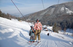Zwei Skifahrer auf einem Schlepplift in einer verschneiten Berglandschaft.