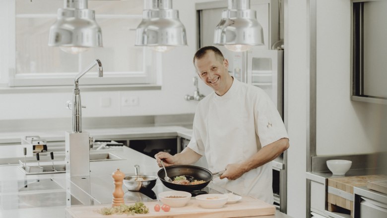 A chef in a modern kitchen holding a pan with a smile.