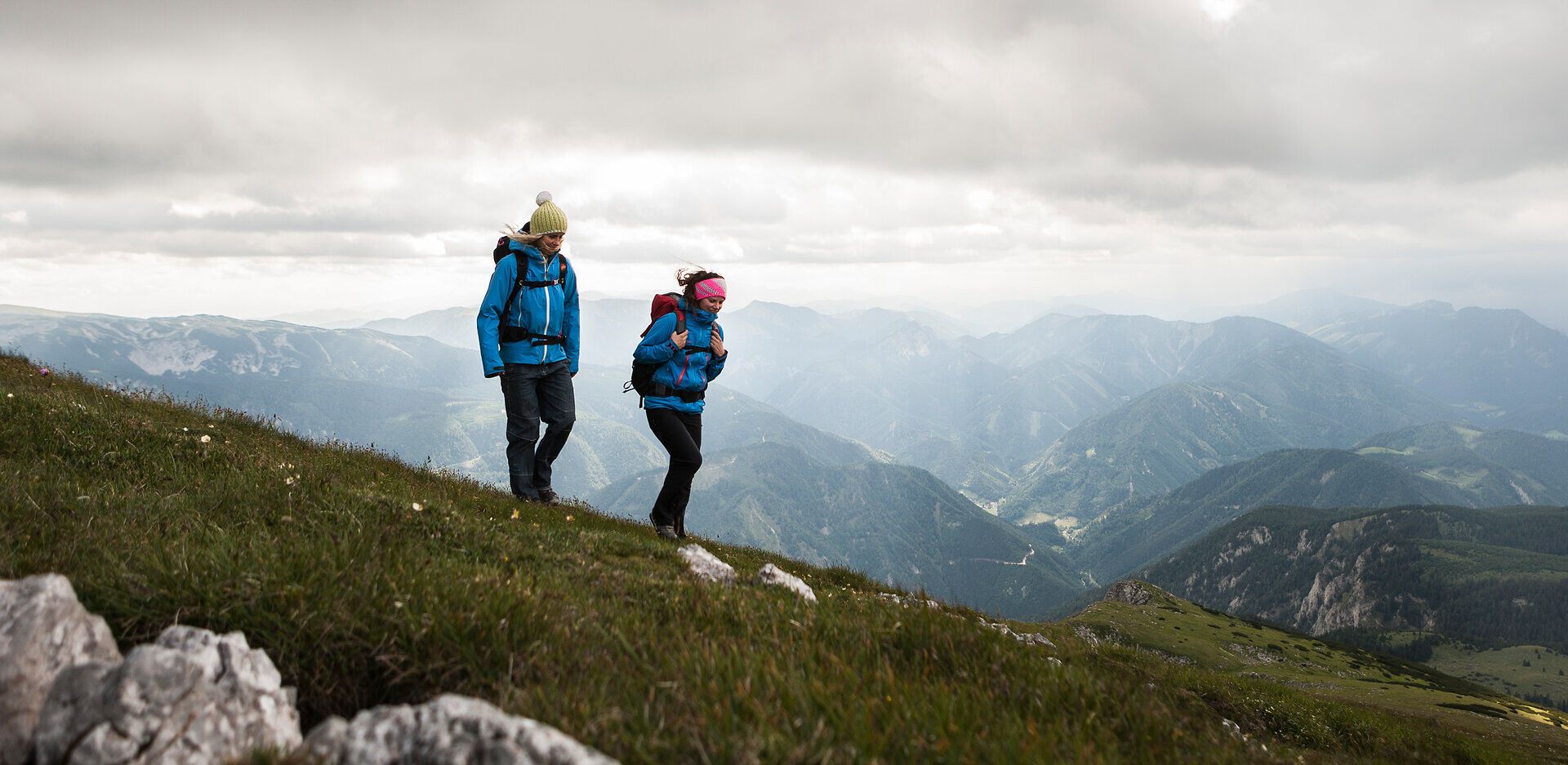 Zwei Wanderer genießen die frische Bergluft und die atemberaubende Aussicht auf die umliegenden Gipfel der Wiener Alpen. Die sanften Hügel und das satte Grün der Wiesen laden zu einem unvergesslichen Abenteuer in der Natur ein. Hier, wo die Freiheit der Berge spürbar ist, wird jeder Schritt zum Erlebnis.