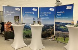 Vor vier Rückwänden einzelner Regionen der Wiener Alpen stehen drei Stehtische, auf denen verschiedene Broschüren angeboten werden. 