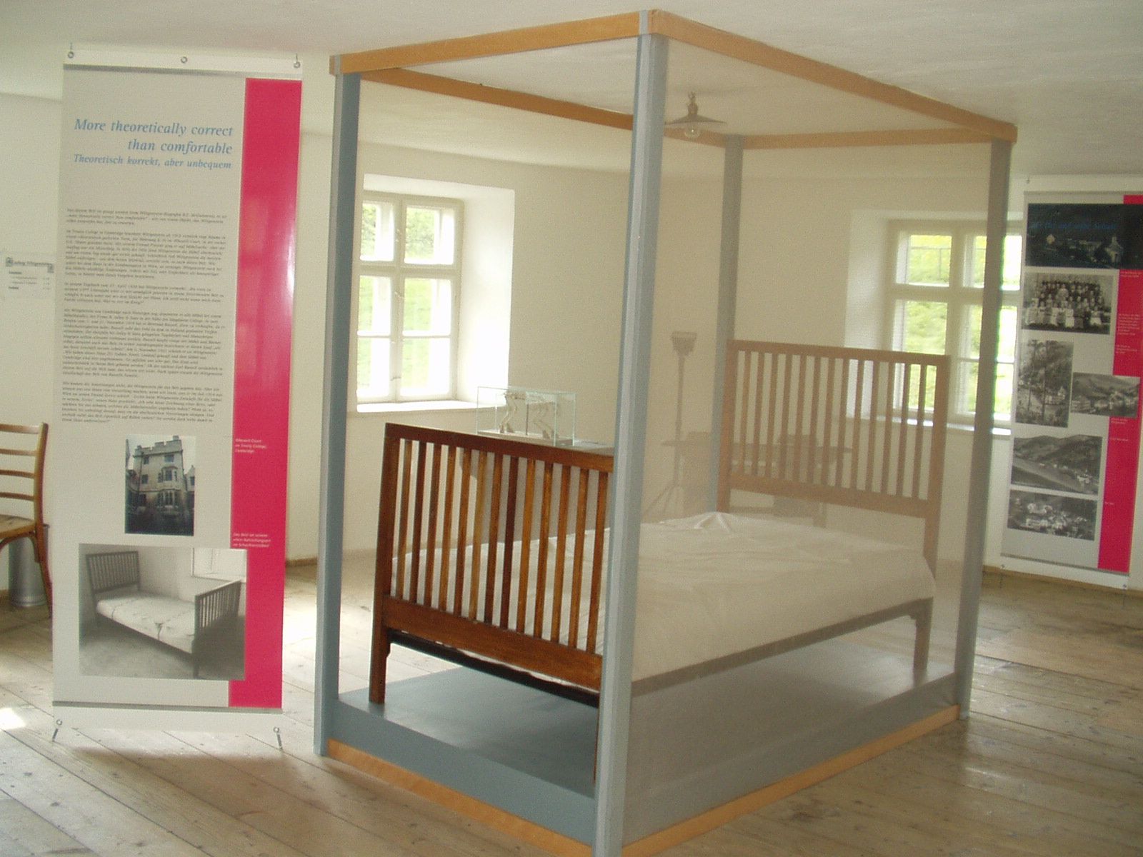 Ausstellungsraum mit einem Bett hinter Glas und Informationstafeln.