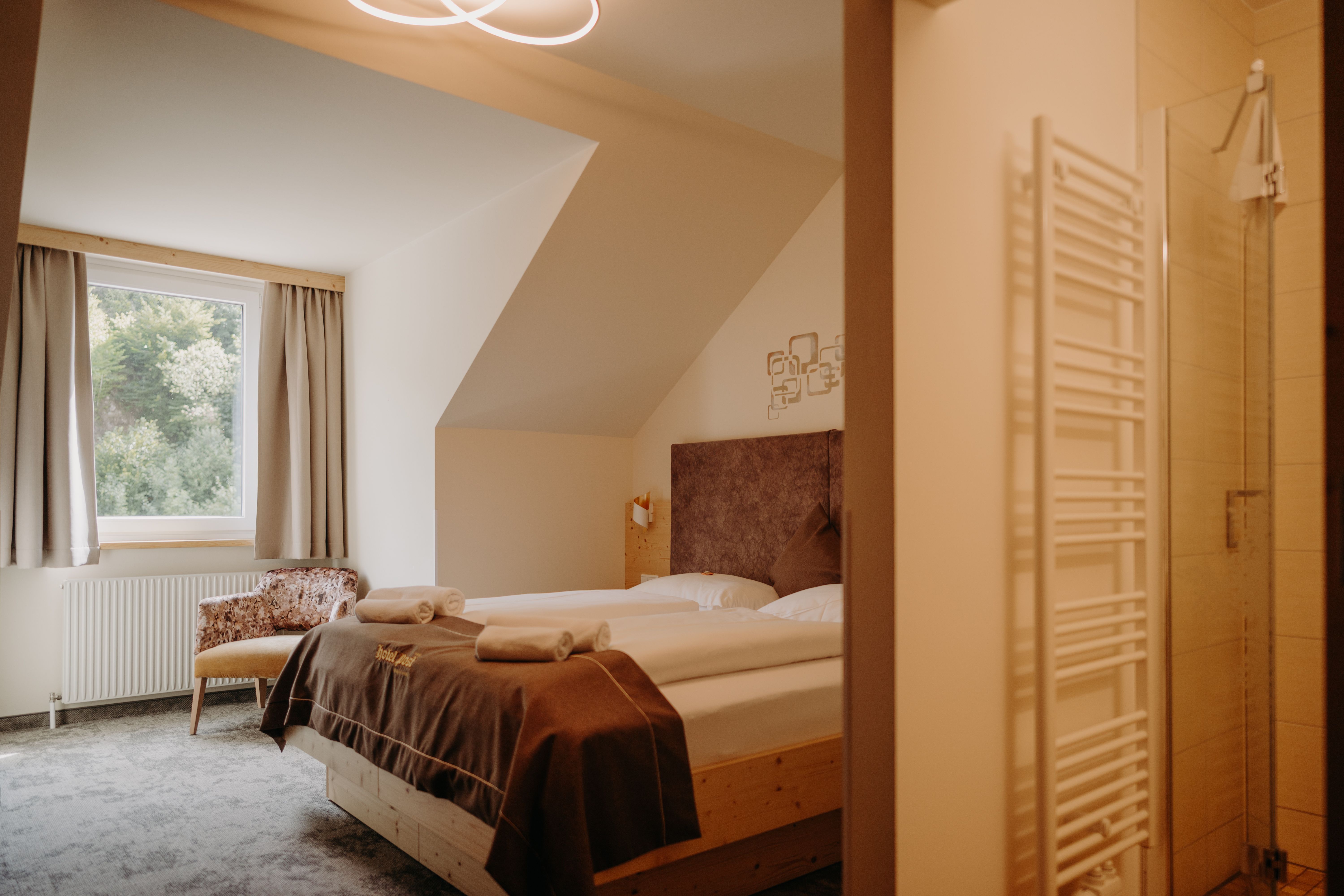 Gemütliches Hotelzimmer mit Doppelbett aus hellem Holz, Fenster mit Vorhängen und einem Sessel. Im Vordergrund sieht man ins Badezimmer. Das Zimmer hat fensterseitig eine Dachschräge.