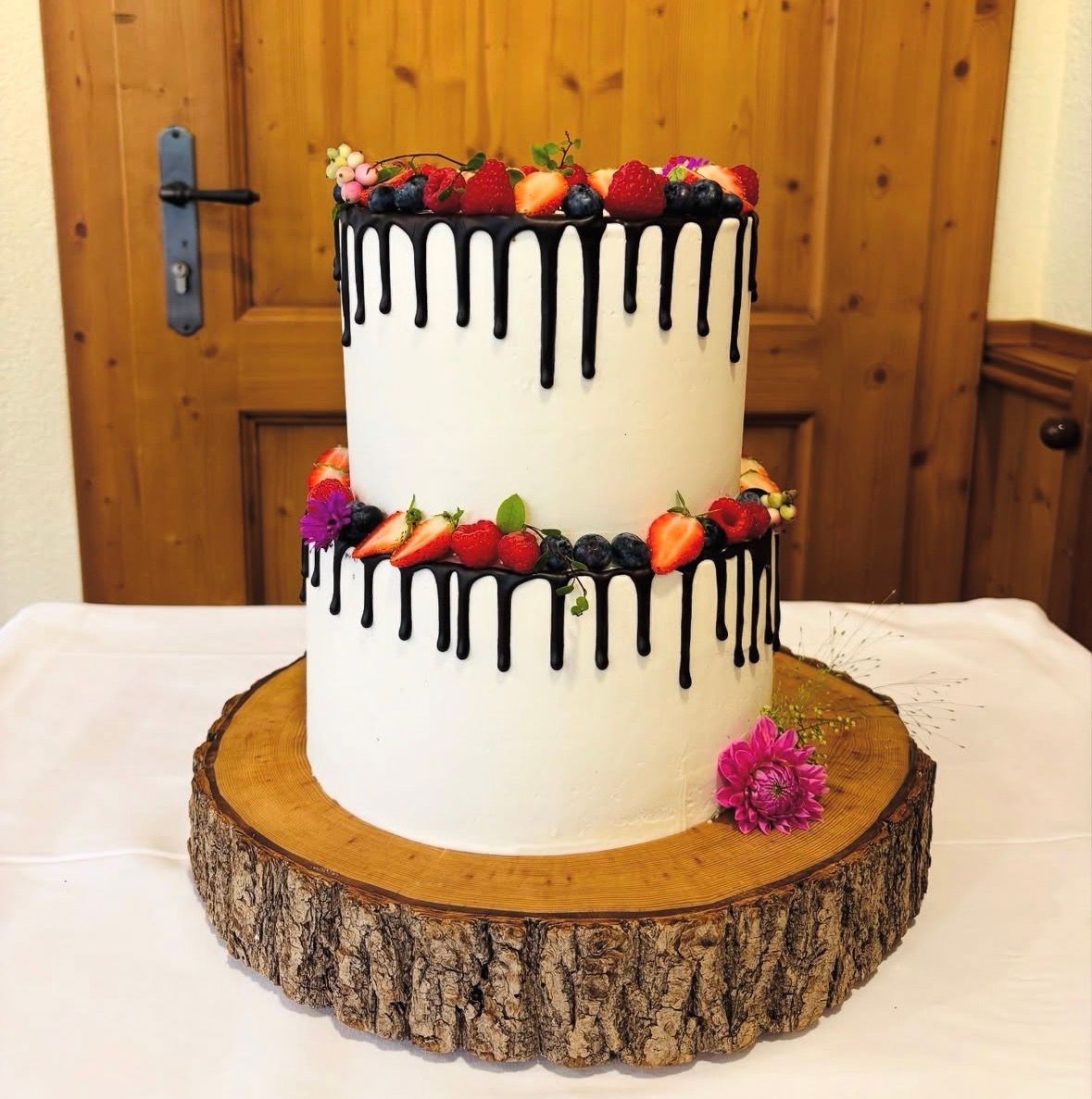 Zweistöckige Torte mit weißer Creme, schwarzem Drip und Beeren auf einer Baumscheibe.