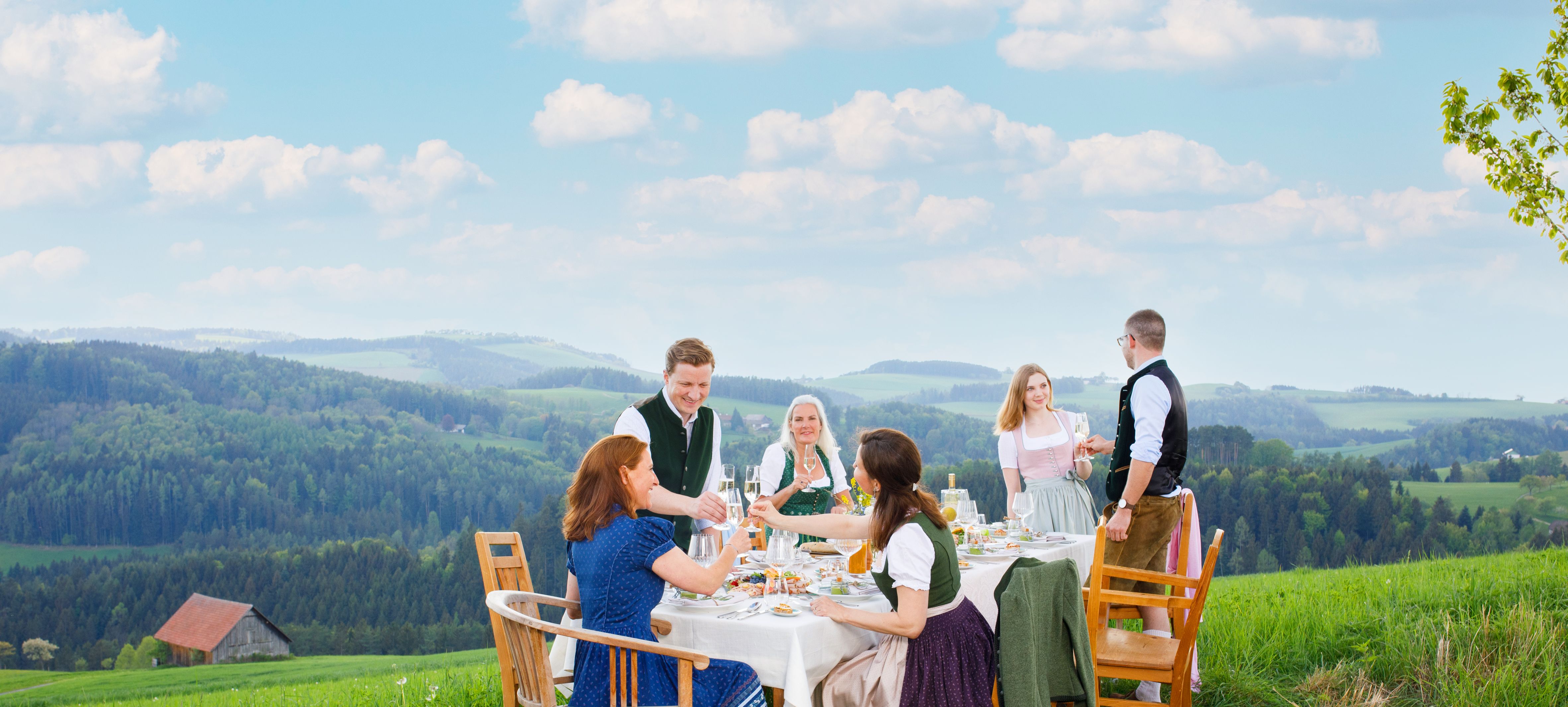 Personen in traditioneller Kleidung rund um gedeckten Tisch im Freien stoßen mit Sektgläsern an.