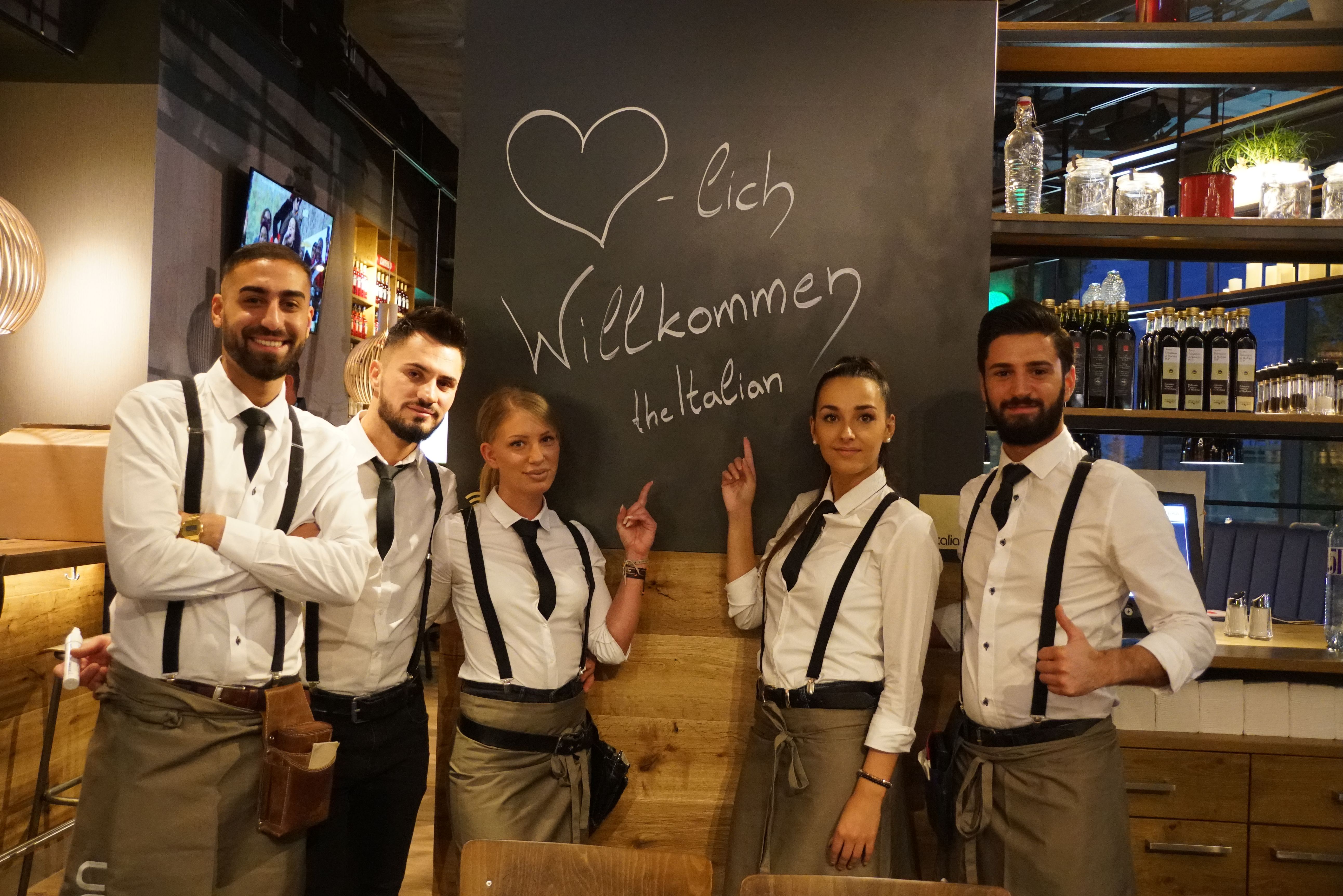 Fünf Personen in Restaurantkleidung stehen vor einer Tafel mit der Aufschrift 'Herzlich Willkommen the Italian'.