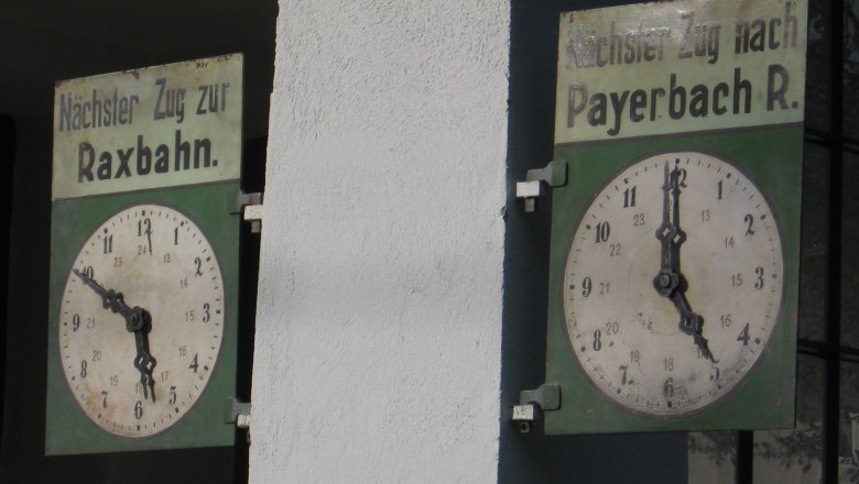 Zwei alte Bahnhofsuhren mit Schildern für Züge zur Raxbahn und nach Payerbach.