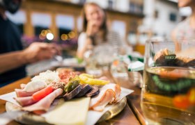 Close-up of a snack with sausage, cheese and vegetables on a table, blurred people in the background.