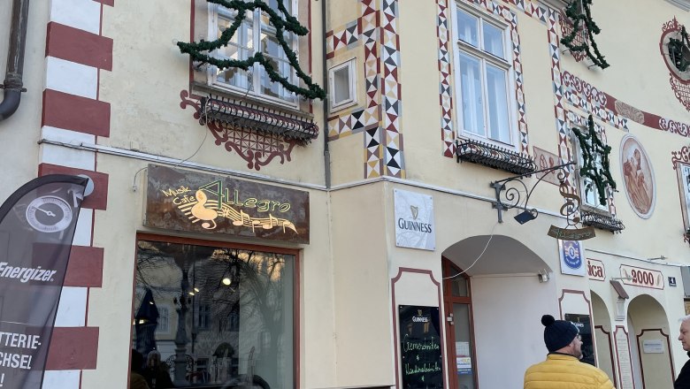 Außenansicht des Cafe Allegro mit dekorierter Fassade und zwei Personen im Gespräch.