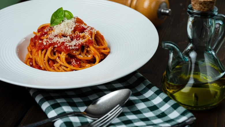 A plate of spaghetti with tomato sauce, basil and parmesan, next to it a bottle of oil and cutlery on a checked cloth.
