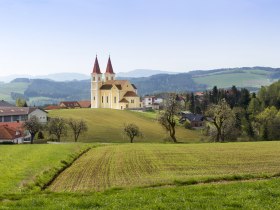 Eine Kirche mit zwei T&uuml;rmen steht auf einem H&uuml;gel in einer l&auml;ndlichen Landschaft mit Feldern und B&auml;umen.