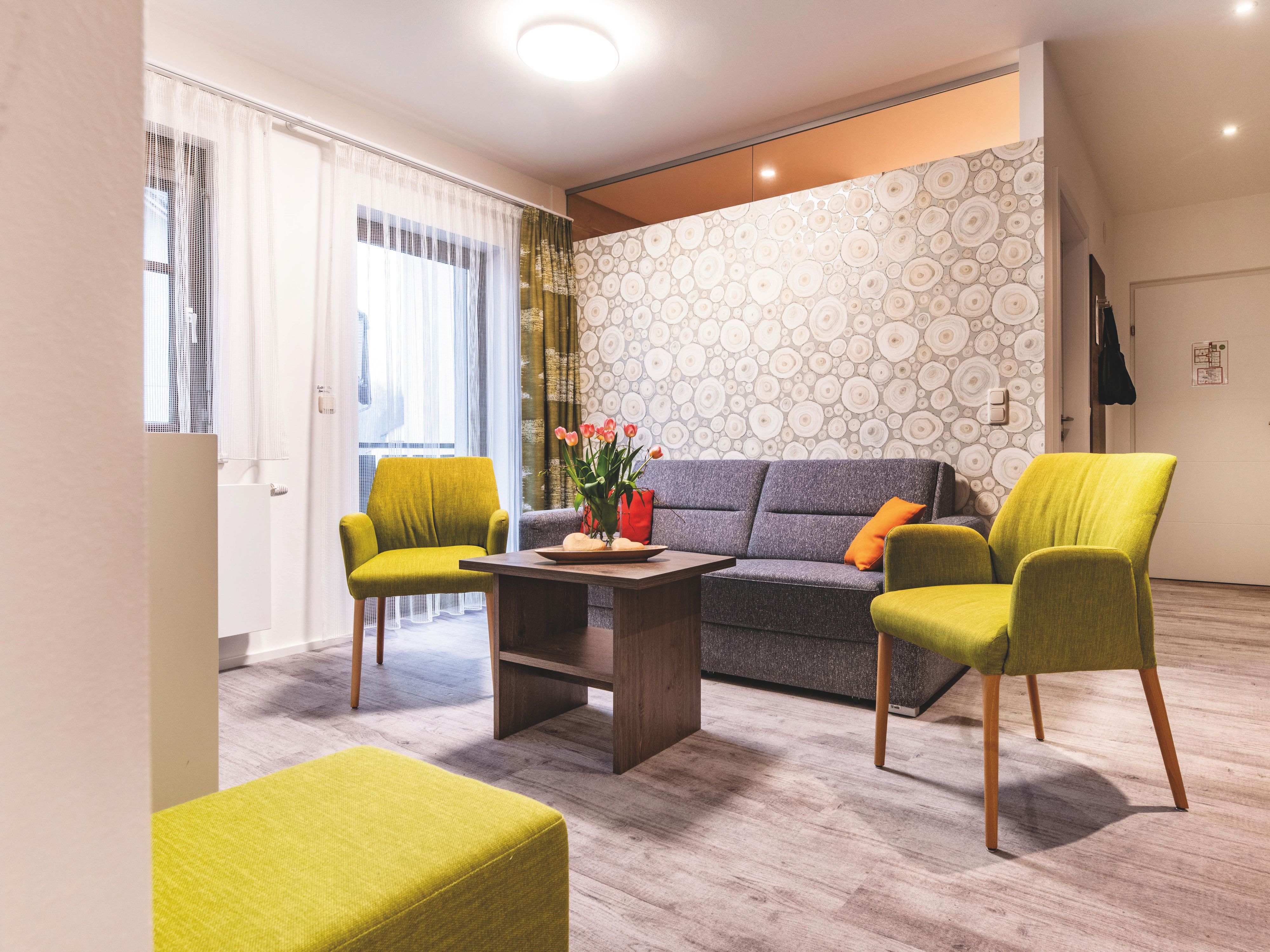 Ein modernes Hotelzimmer mit Couch, zwei Stühlen einem Couchtisch und Fenstern mit Vorhängen.