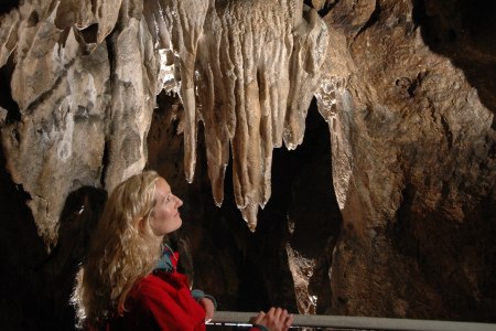 Eine Frau in roter Jacke betrachtet Stalaktiten in einer H&ouml;hle.