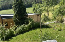 Ein modernes Holzhaus in einer gr&uuml;nen, h&uuml;geligen Landschaft mit B&auml;umen und Wiesen.
